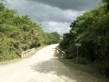 A quiet, unpaved rural road winds through dense green vegetation under a cloudy sky, featuring a small bridge and a roadside sign.