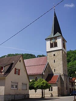 A stone church tower with a clock stands beside traditional village houses with red-tiled roofs under a clear blue sky in Lenningen.