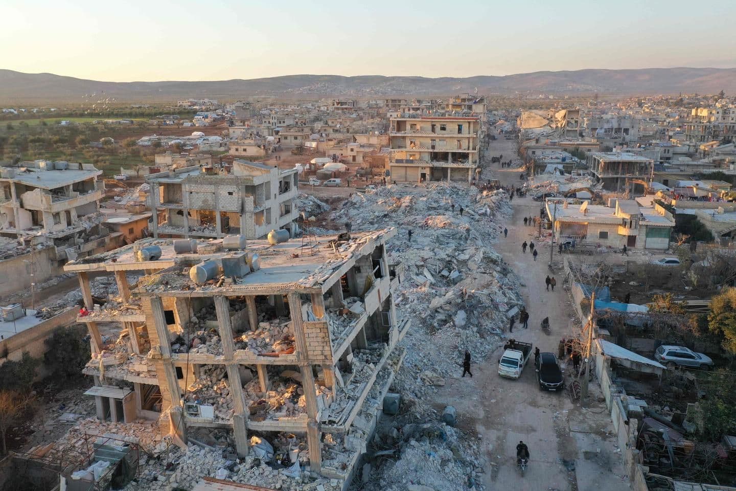 Aerial view of a war-damaged town with rubble-filled streets, ruined buildings, and scattered vehicles under a clear sky.