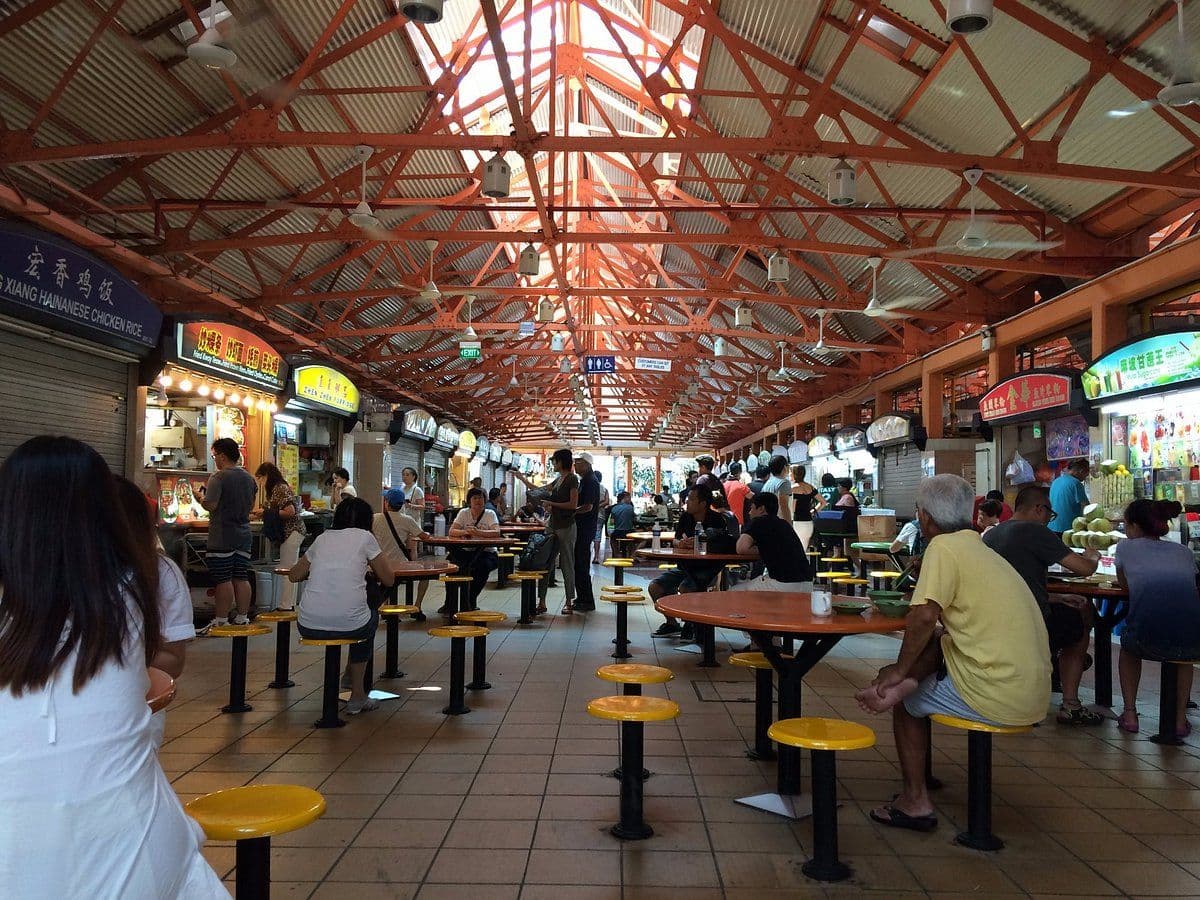 Interior view of a bustling Singapore hawker centre featuring orange steel trusses, food stalls, and diners at tables.