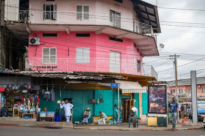A colorful pink and teal building with street-level shops, pedestrians, and overhead wires in a town center streetscape.