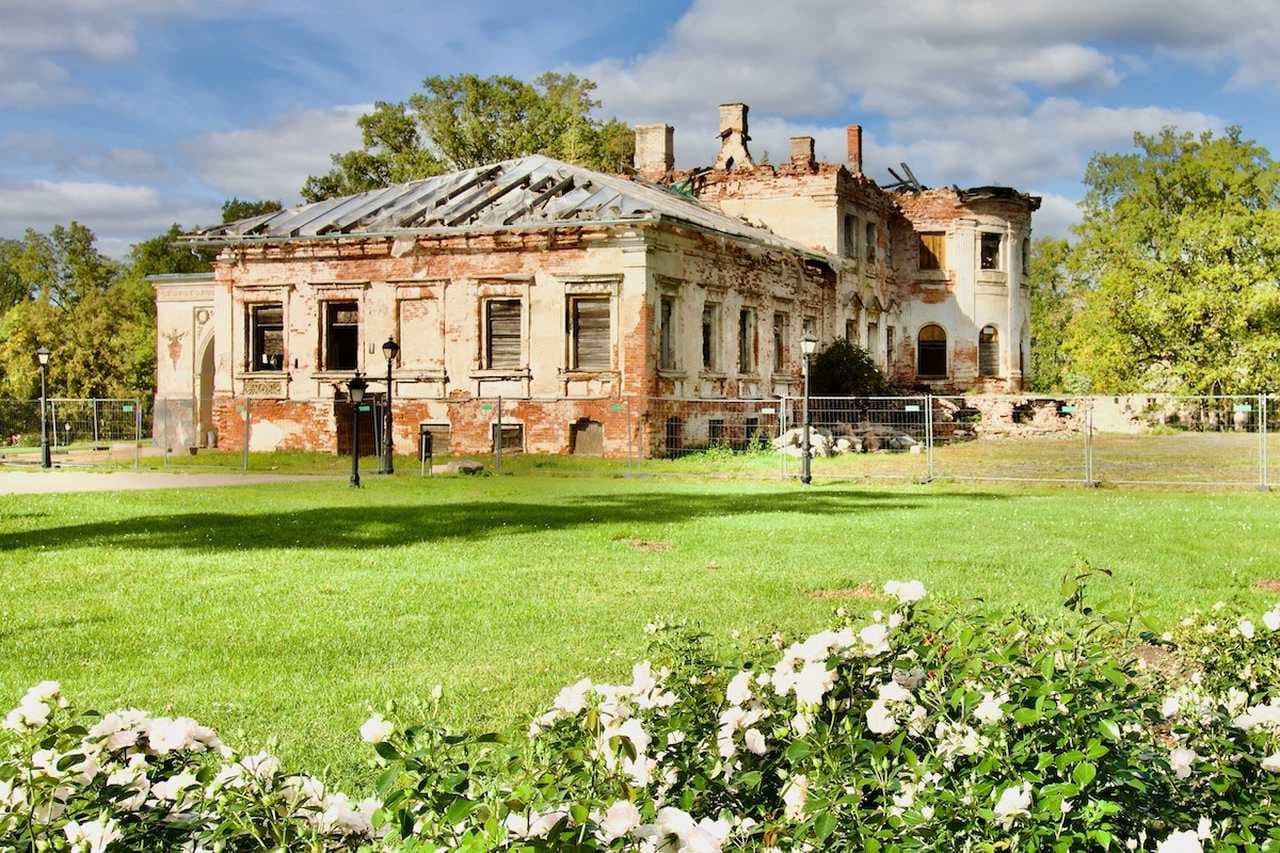 Ruins of a historic manor house in Vidzeme, Latvia, surrounded by green lawns and white flowers under a blue sky.