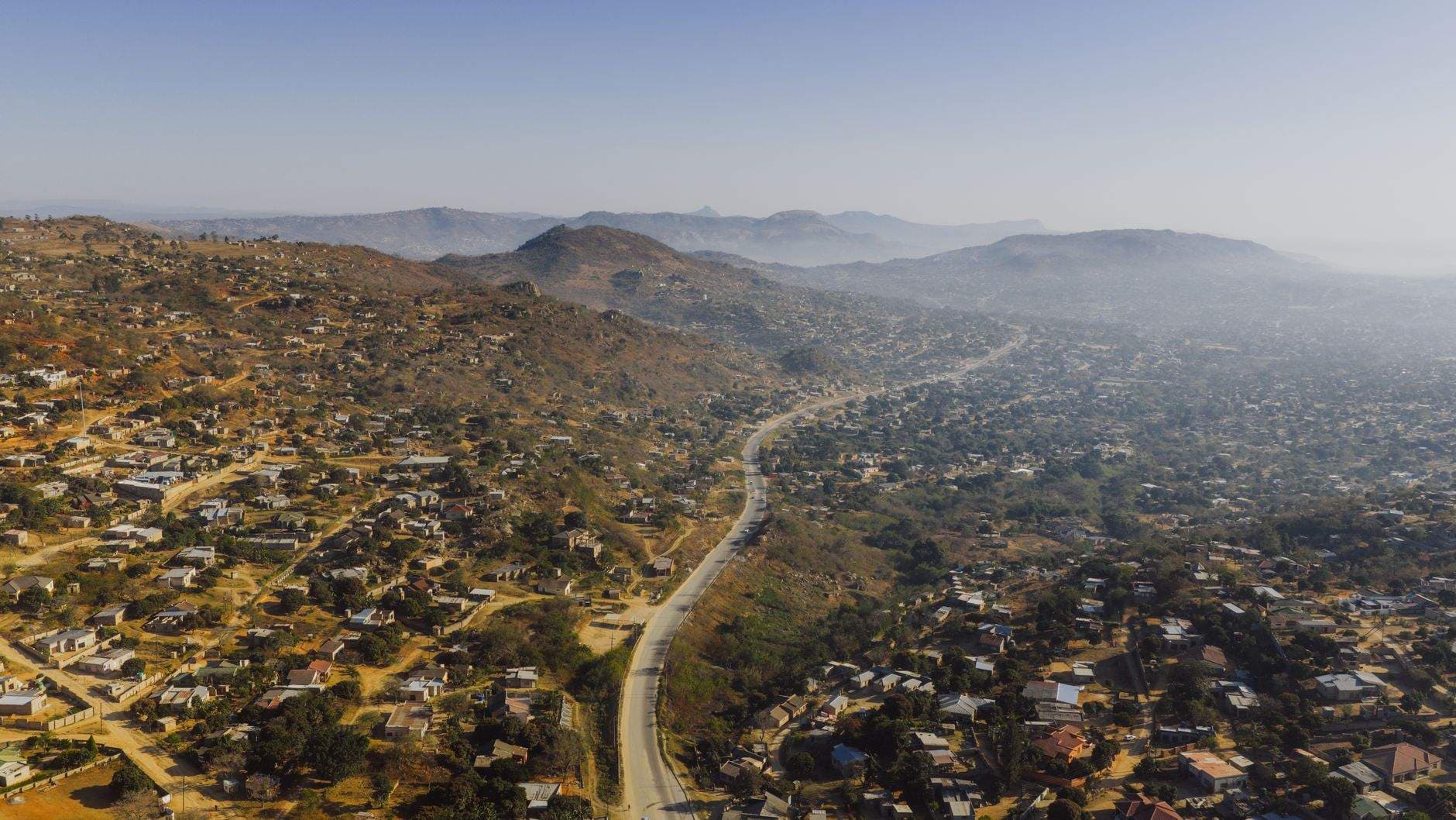 Aerial view of a winding road cutting through a rugged, hilly landscape dotted with scattered houses and vegetation under a hazy sky.