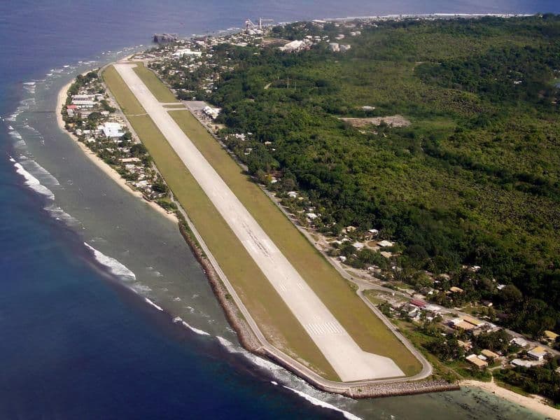 Aerial view of a long coastal airstrip on a narrow island, with ocean waves on one side and dense green vegetation plus scattered buildings inland.