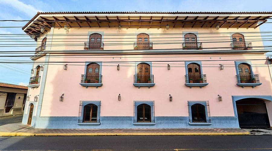 A pink colonial-style building with blue trim and wooden balconies stands on a street corner under a blue sky.