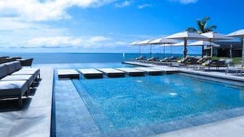 A luxury resort infinity pool overlooking the ocean, featuring lounge chairs, white umbrellas, and a clear blue sky.