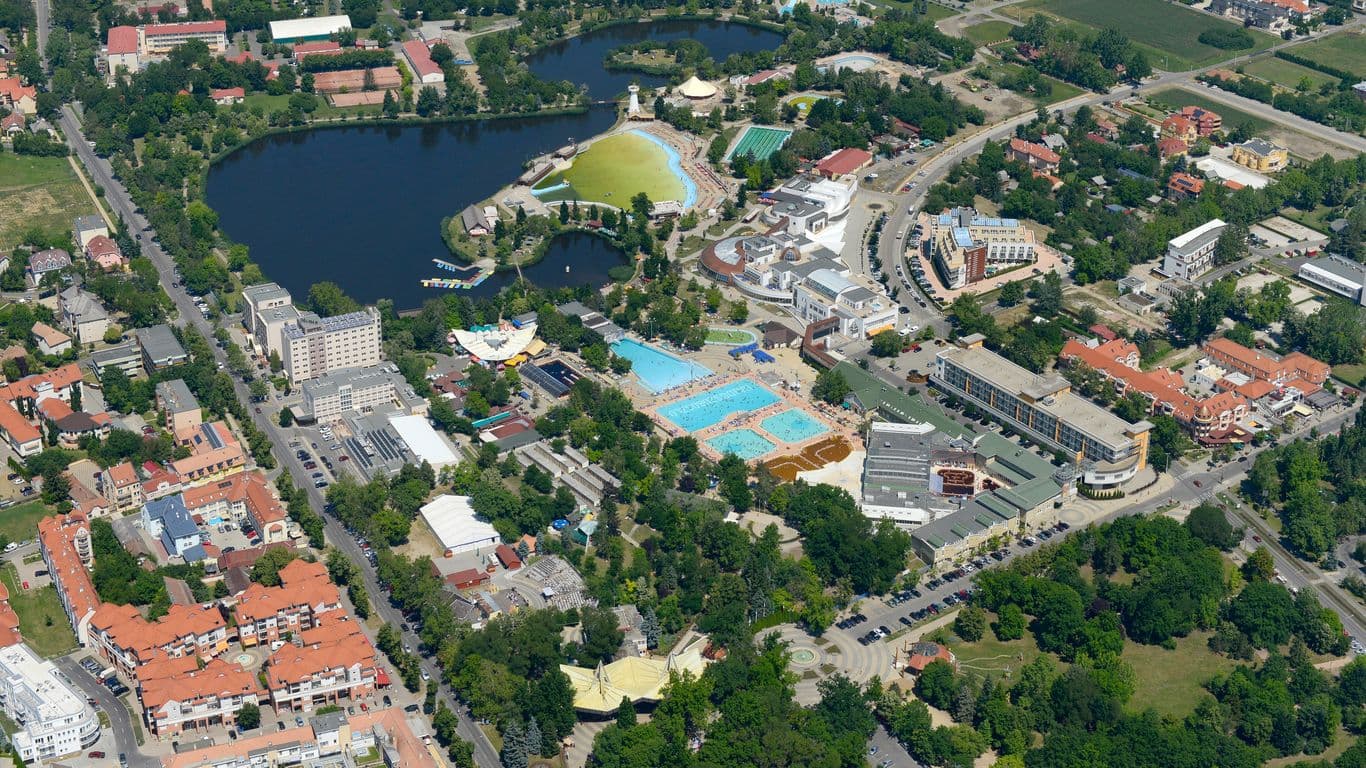 Aerial view of a resort complex in Vecsés featuring swimming pools, a large lake, and surrounding residential buildings.