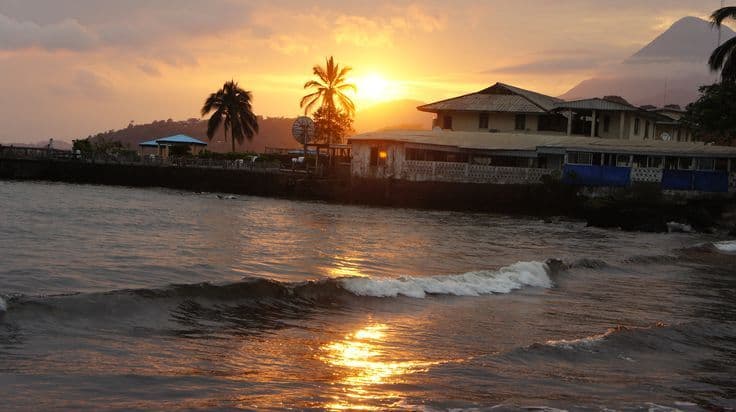 Coastal sunset scene with waves, palm trees, and a waterfront building in Limbe, Cameroon, under a golden sky