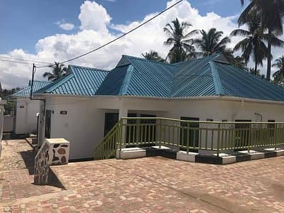 Exterior view of a white single-story building with a blue metal roof and green railing, surrounded by palm trees and a paved courtyard.
