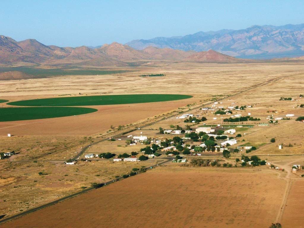Aerial view of a dry highland valley with arid fields, a small settlement, and distant mountains under a clear blue sky.