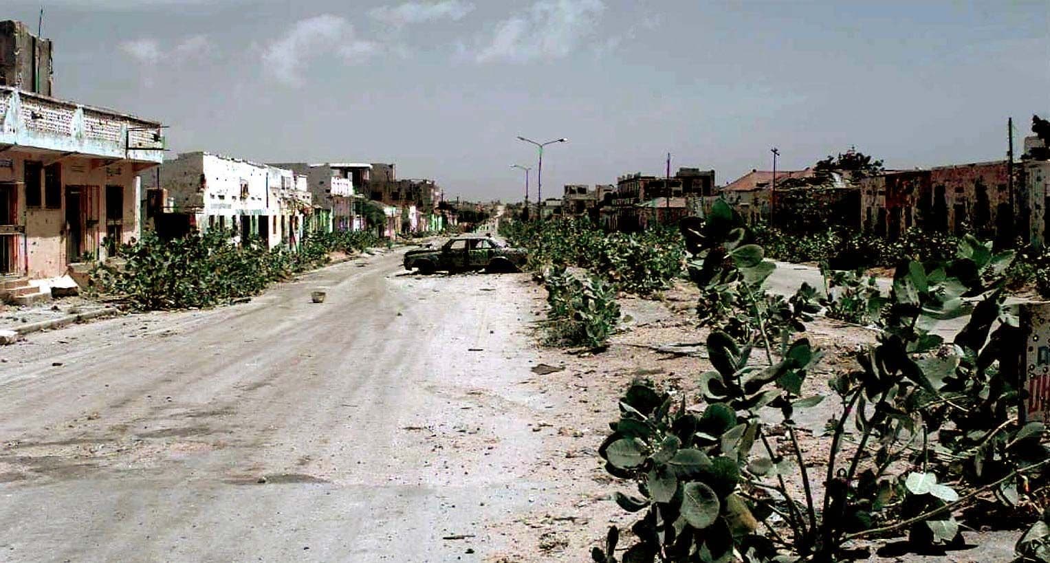 Dusty unpaved road through a small town with low buildings, overgrown vegetation, and a parked car under a hazy sky.
