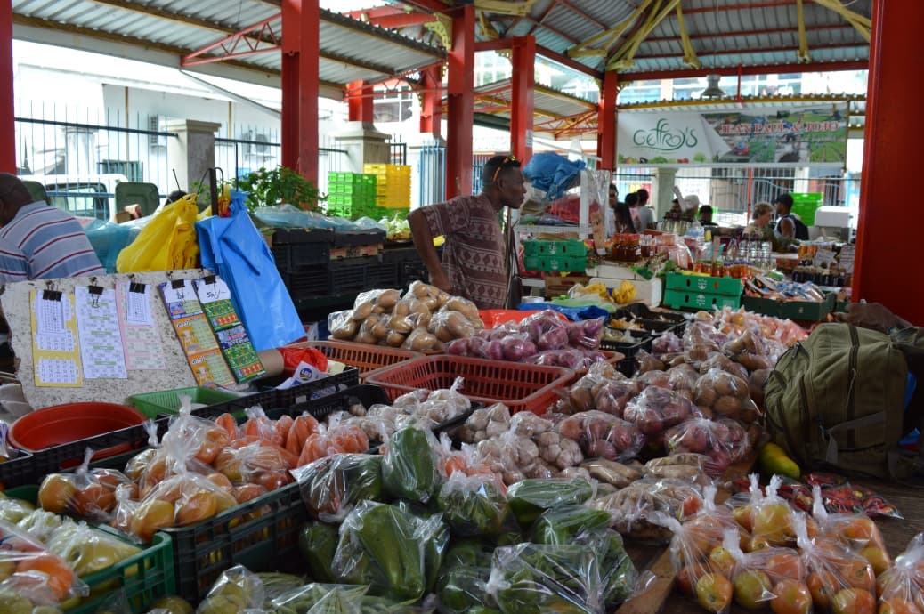 A bustling market stall displays various bagged produce like potatoes, peppers, and onions under a red-pillared shelter with vendors in the background.