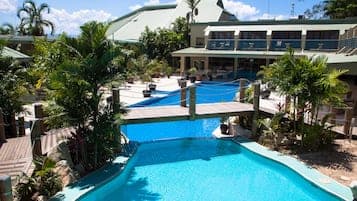 Aerial view of a resort complex featuring a large swimming pool, lush tropical vegetation, and multi-story guesthouse buildings under a bright sky.