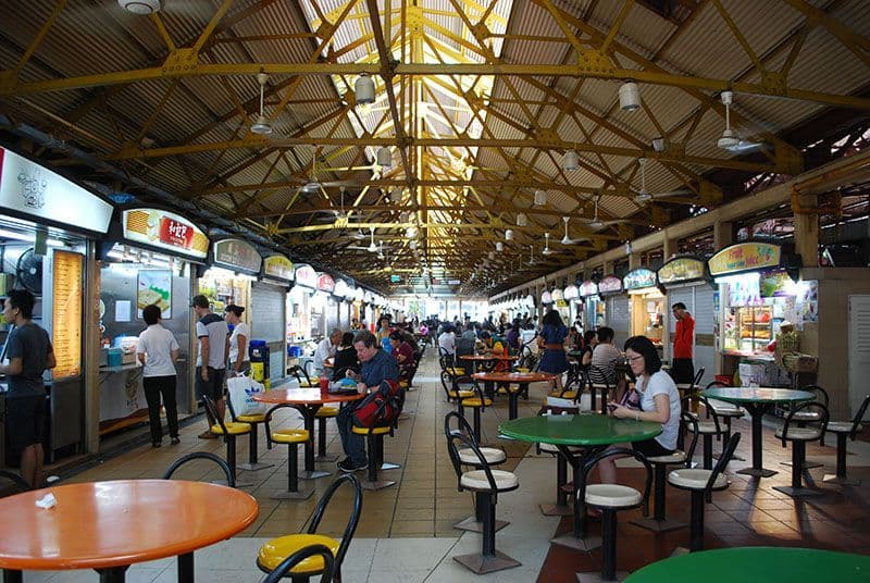 Interior view of a bustling Singapore hawker centre featuring rows of food stalls, colorful tables, and diners under a yellow steel roof.