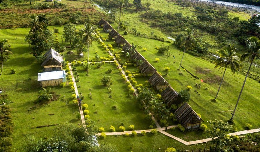 Aerial view of a row of thatched-roof eco-lodge bungalows situated on a lush green lawn with palm trees and winding pathways at Namosi Eco Retreat.