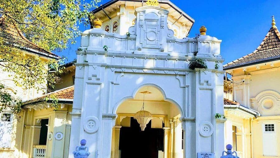 Sunlit white arched gateway of Asokaramaya Buddhist Temple in Thimbirigasyaya, featuring ornate detailing and flanking yellow buildings under a clear blue sky.