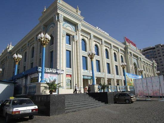 Street view of a large, ornate building with blue accents and Cyrillic signage in Bokhtar, featuring parked cars and decorative lamp posts.