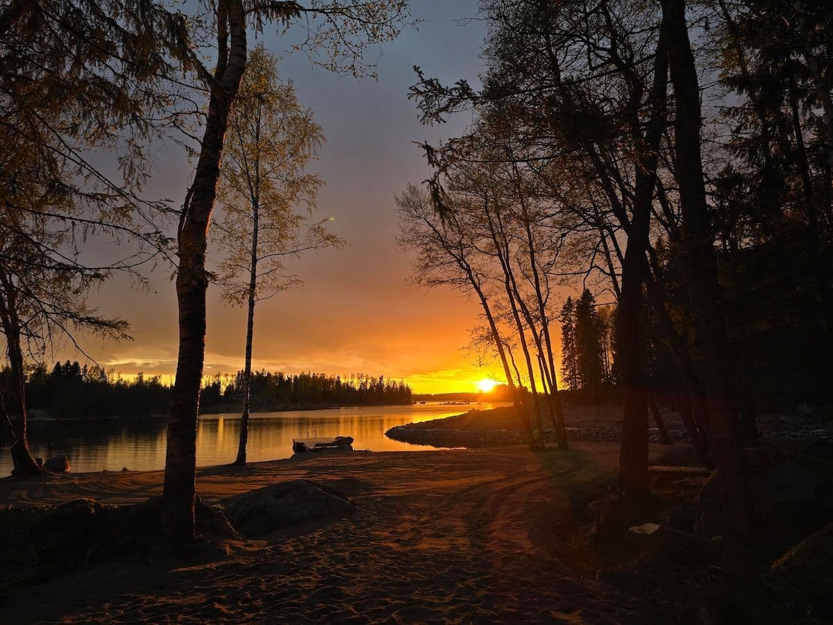 Sunset over a calm lake shoreline in Kaustinen, featuring silhouetted trees, a sandy beach, and a small boat.