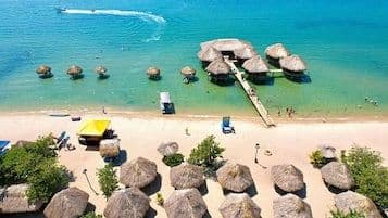 Aerial view of a tropical beach resort with thatched-roof huts on land and over turquoise water.