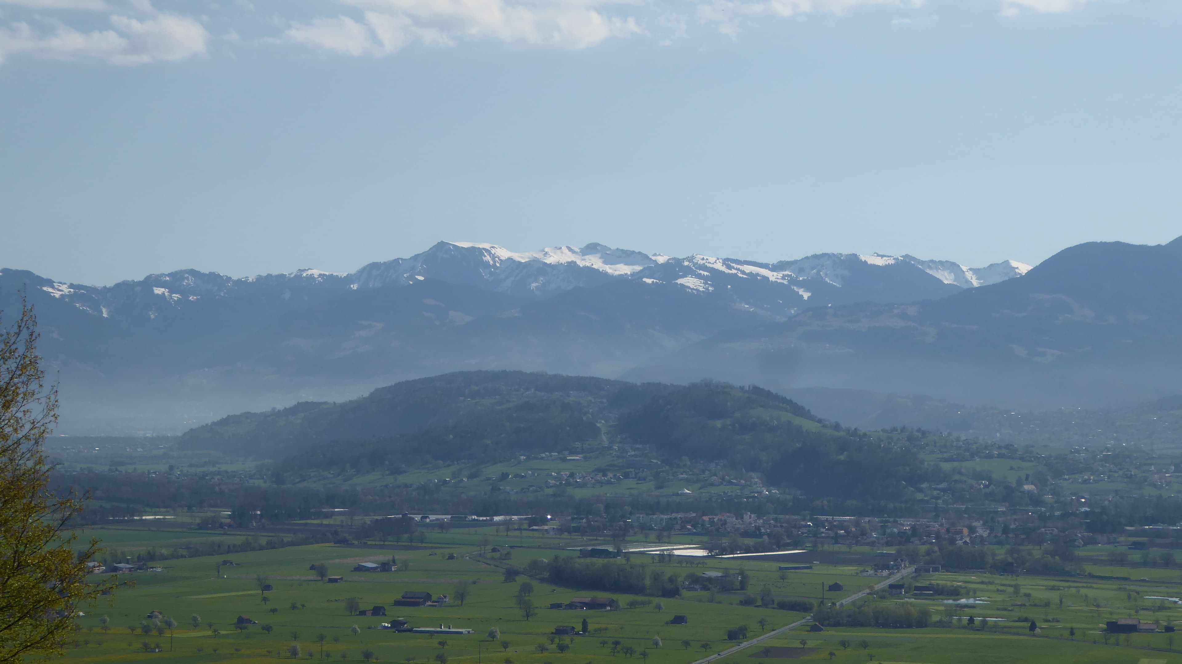 Wide landscape view of a green valley with scattered houses and distant snow-capped mountains under a blue sky.