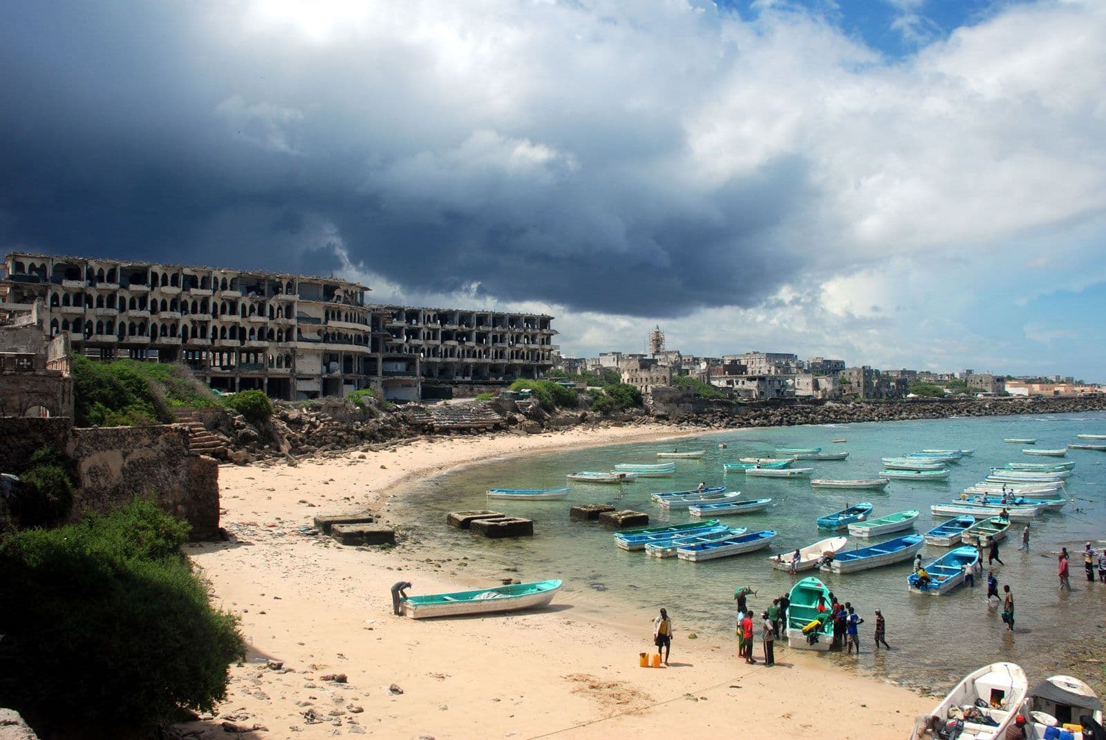 Coastal shoreline with sandy beach, numerous small boats in calm water, ruined multi-story buildings, and scattered people near boats under dramatic storm clouds.
