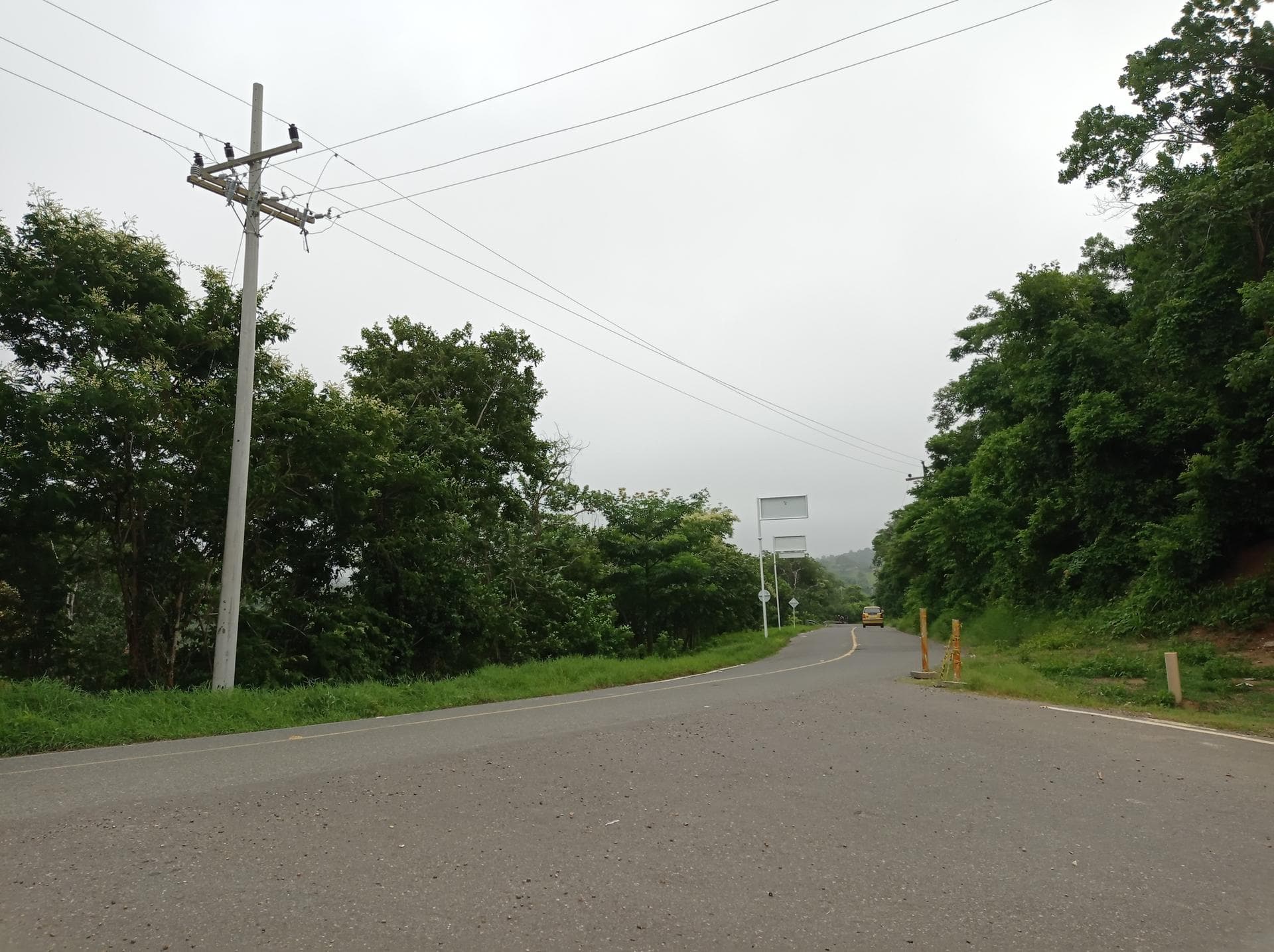 A paved rural road winds through lush green vegetation under an overcast sky, featuring a utility pole and a distant yellow vehicle.