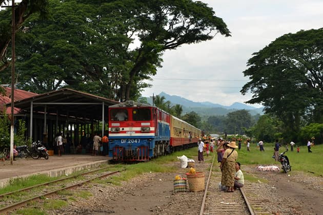 A colorful Myanmar train arrives at a rural station near Mandalay, with passengers and vendors waiting on the tracks under large trees.