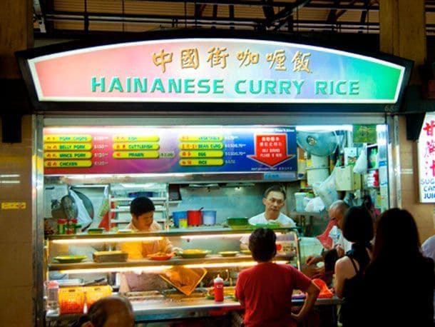 A brightly lit Hainanese Curry Rice stall in a hawker center, showing vendors preparing food and customers waiting at the counter.