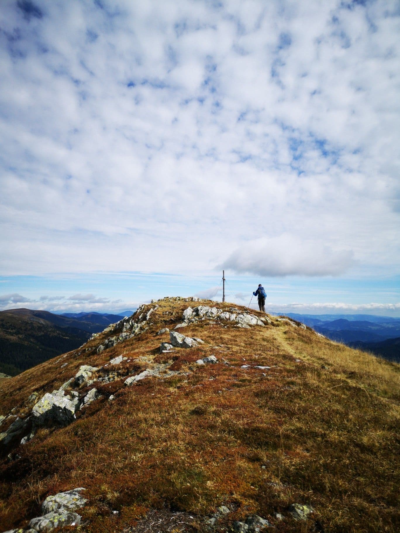 A lone hiker stands on a rocky, grassy mountain ridge under a vast sky filled with scattered clouds, overlooking distant mountain ranges.