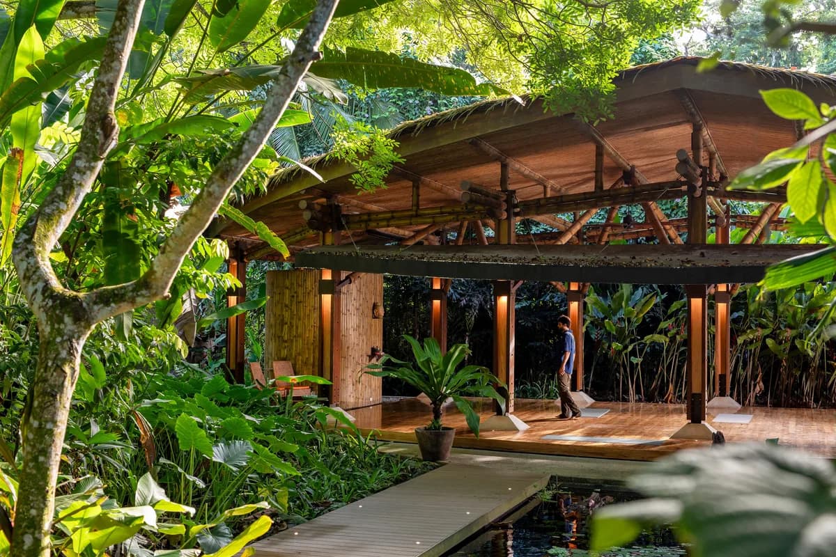 Open-air bamboo and wood yoga shala with thatched roof, surrounded by lush tropical jungle foliage and a small water feature.