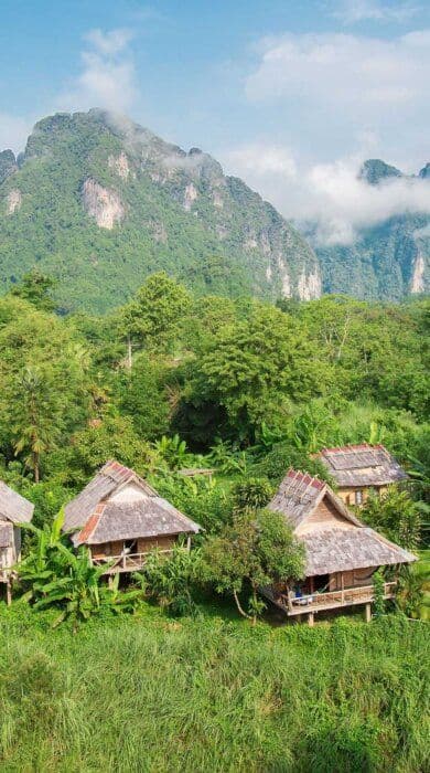 Traditional wooden stilt houses with thatched roofs sit in a lush green valley, framed by towering limestone mountains and misty clouds.
