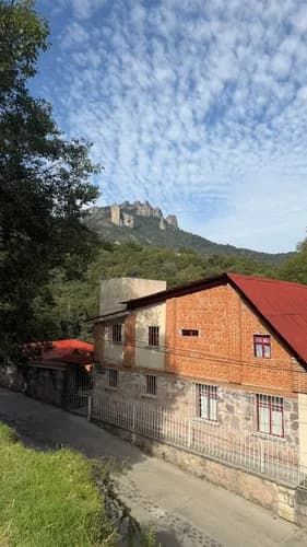 A rural village street with brick and stone houses, a red roof, and a hillside mountain backdrop under a cloudy sky.