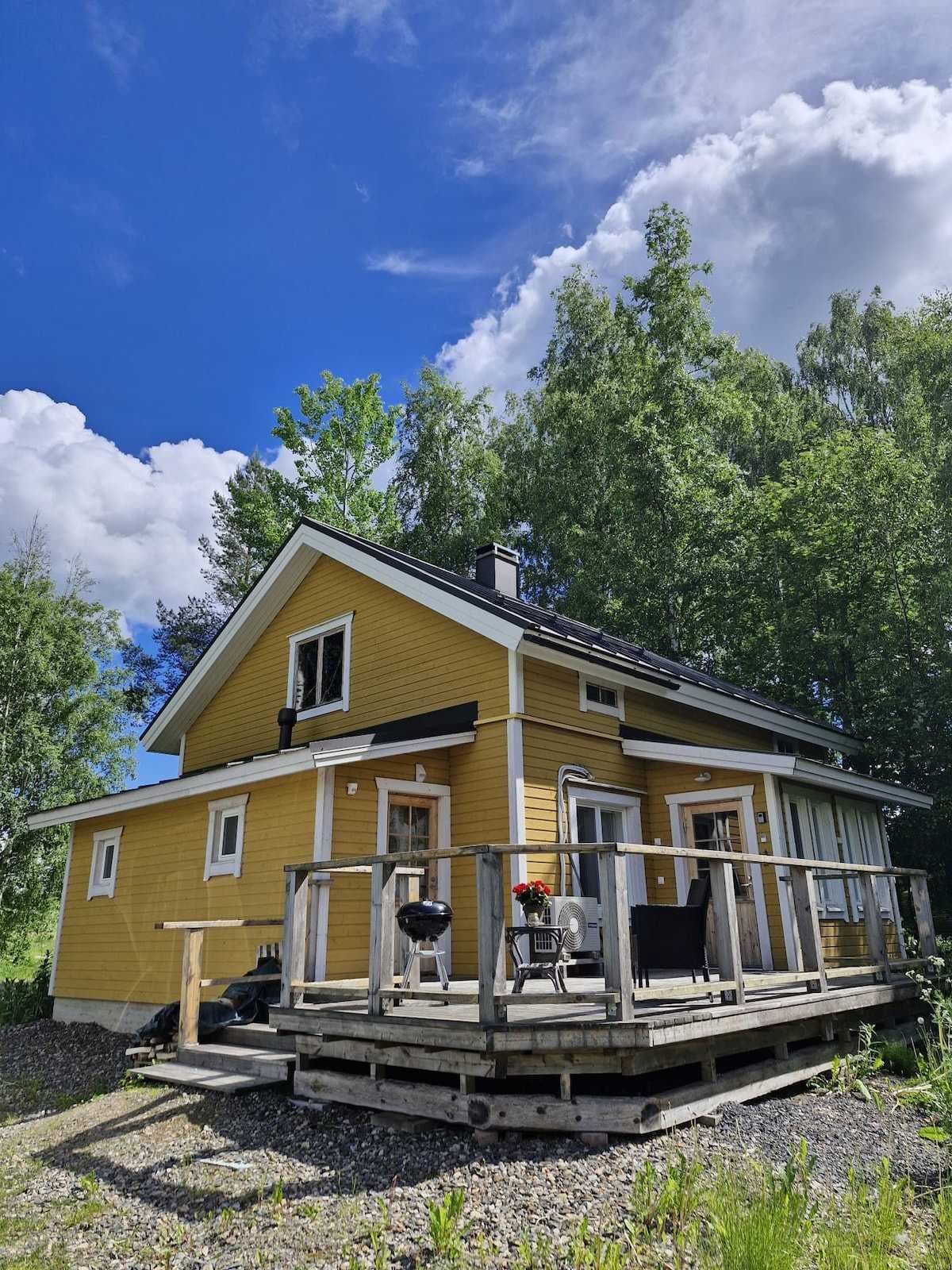 A yellow traditional wooden cottage with a wooden deck and grill, surrounded by green trees under a blue sky with clouds in the Kaustinen region.