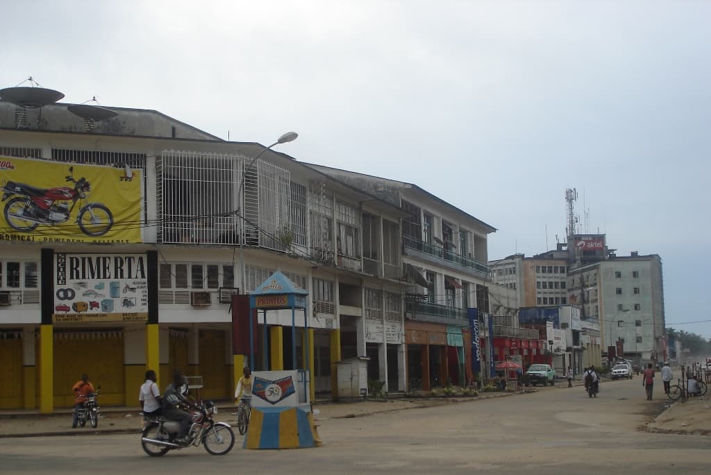 Street level view of Kisangani showing commercial buildings, a motorcycle billboard, and daily traffic activity under an overcast sky.