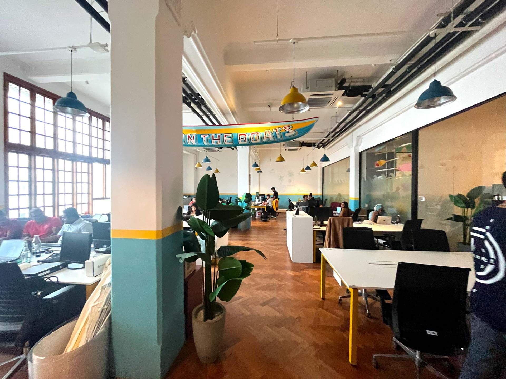 Interior view of a modern co-working space featuring wooden floors, colorful pendant lights, desks with laptops, and a decorative boat sign.