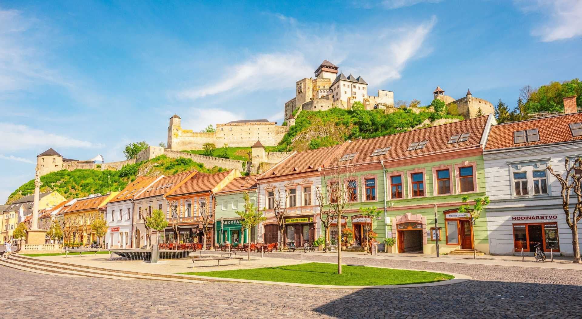 Wide street scene of Bánovce nad Bebravou town centre with colorful shops, outdoor cafe seating, and the hilltop castle in the background.