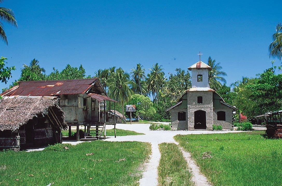 Rural West African village scene with a stone church, thatched huts, palm trees, and a dirt path under a clear blue sky.