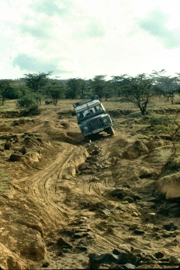A 4x4 vehicle navigates a rough dirt track through a dry, rocky landscape with scattered acacia trees under a cloudy sky.