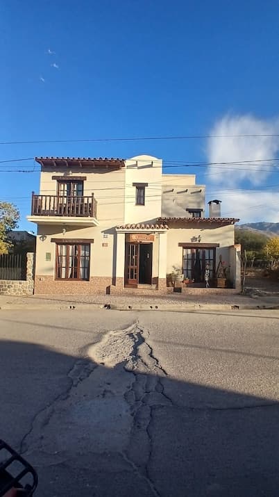 Street view of a beige two-story house with a wooden balcony and tiled roof in Simoca, set against a blue sky with distant mountains.