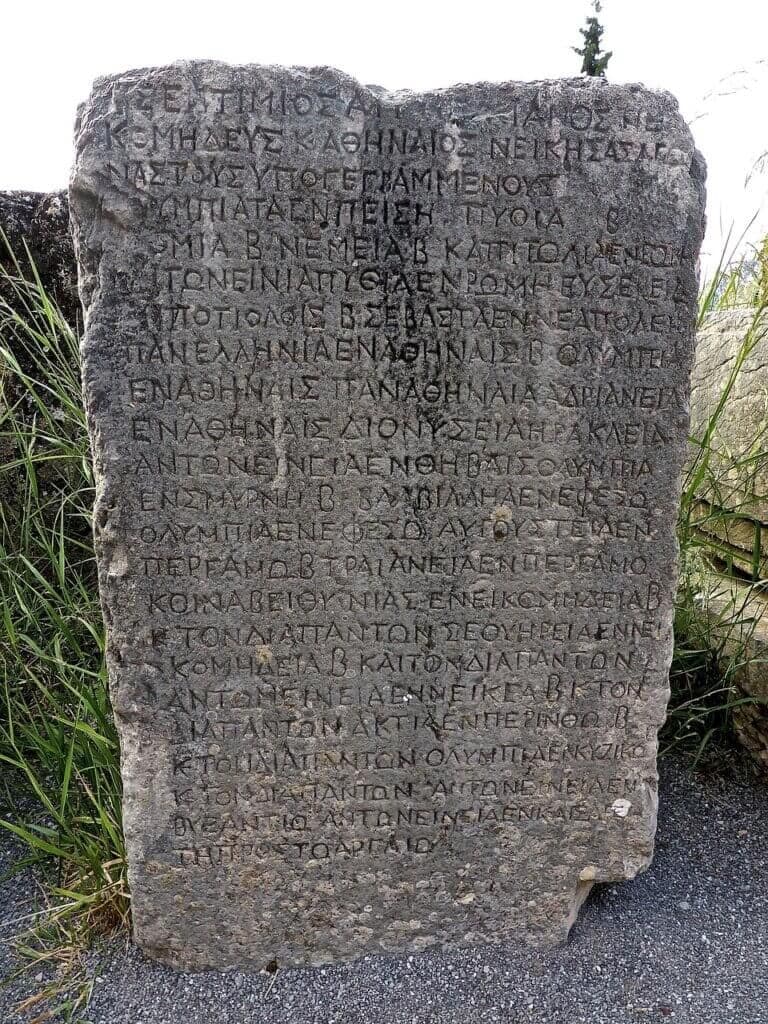A large, weathered stone stele with Greek inscriptions stands outdoors on gravel, surrounded by grass.