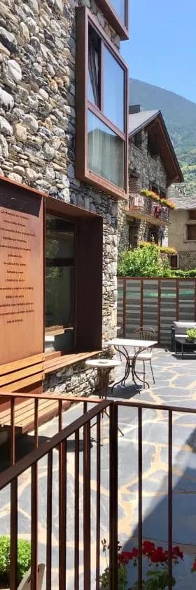 Stone building exterior with wooden window frames and patio furniture in Ramio, Andorra, featuring a menu board and mountain backdrop.
