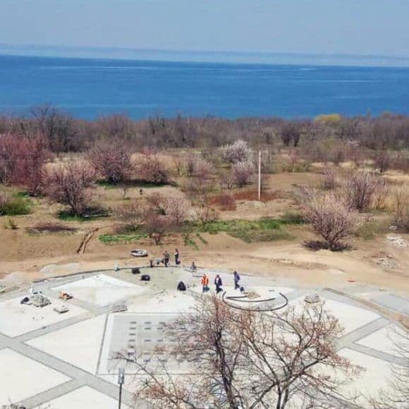 Aerial view of a paved public plaza in Dniprorudne with scattered people, adjacent park trees, and a large body of water.