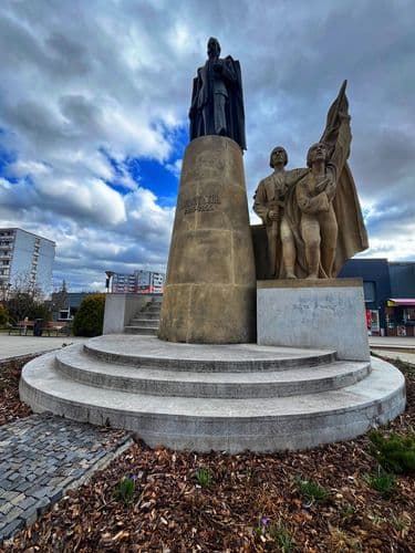 Bronze WWII memorial monument with a standing figure and two soldiers holding a flag, set in a paved town square in Bánovce nad Bebravou.