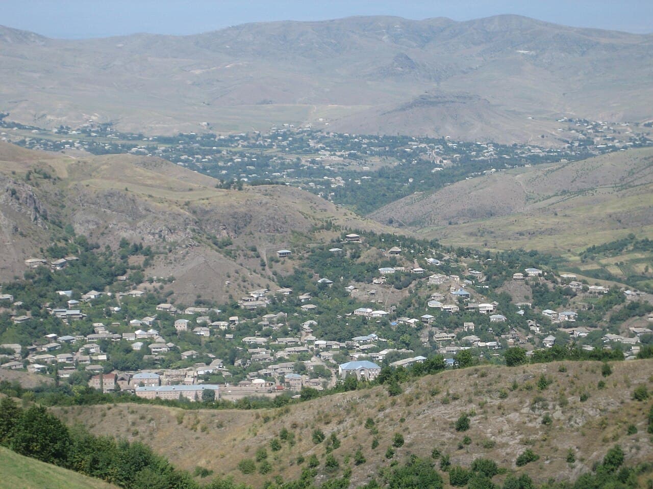 Aerial view of a village nestled in a valley with rolling hills and mountains in the background.
