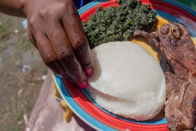 A hand reaches toward a plate of white sadza, green vegetable relish, and grilled roadrunner meat in Marondera, Zimbabwe