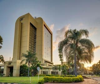 Exterior view of a large beige hotel building with palm trees, manicured lawn, and a sunset sky.