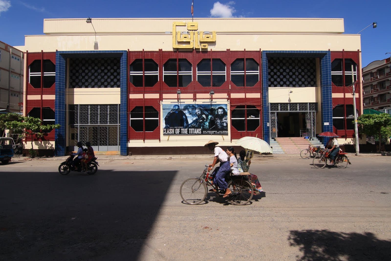 A street scene in Mandalay featuring a cinema with a Clash of the Titans poster, while a cyclo and motorbikes pass by on the road.