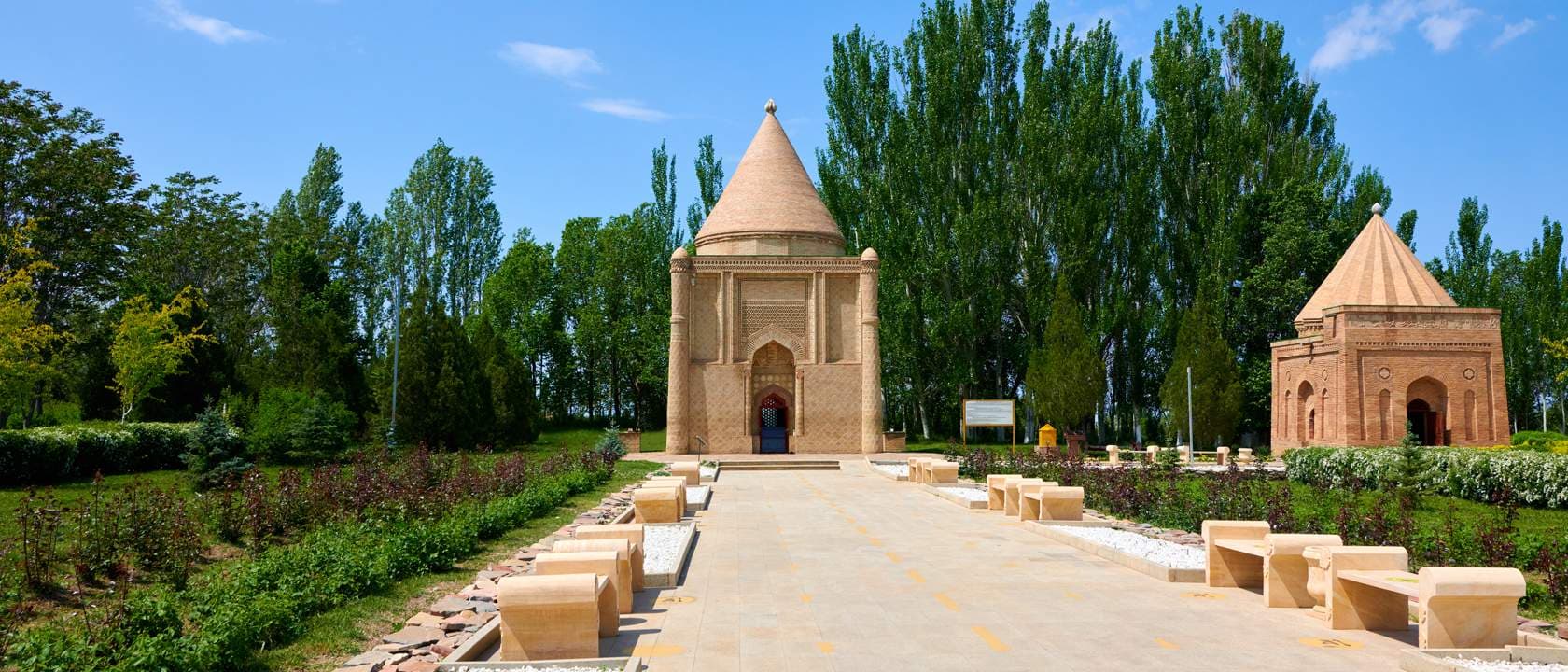 Exterior view of the Aisha Bibi mausoleum in Taraz, featuring a conical brick dome, paved walkway, stone benches, and surrounding greenery under a clear blue sk