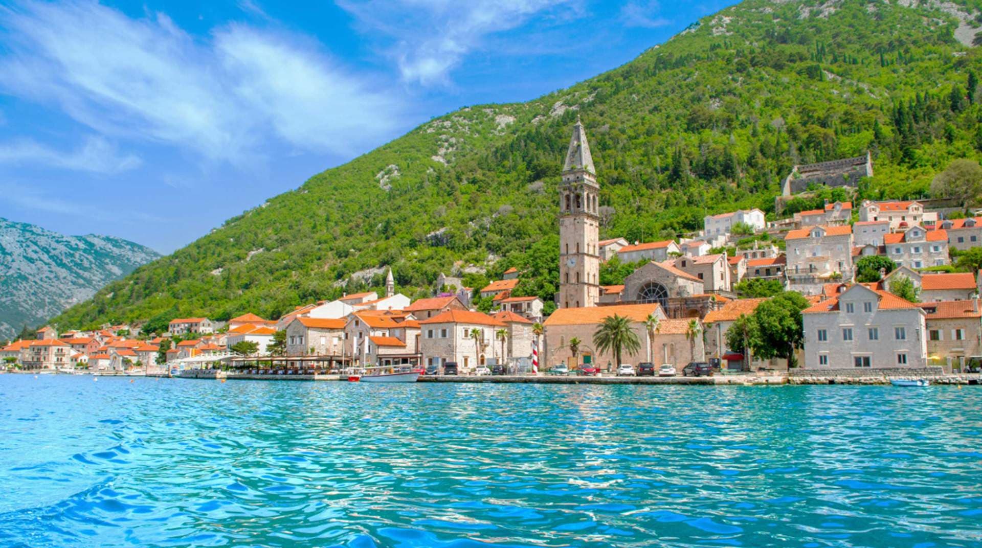 Coastal view of Tivat Old Town with red-roofed buildings, a prominent bell tower, and turquoise water in the foreground.