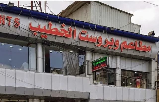 Exterior view of a commercial building facade featuring large red Arabic signage and English text, typical of a street scene in Ash Shaghadirah.
